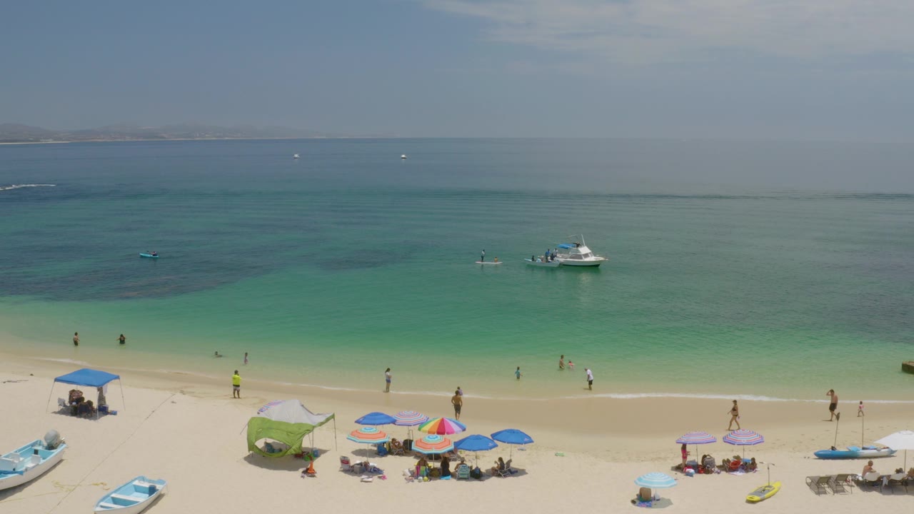 Aerial view of Palmilla Beach in Baja California Sur with umbrellas on pale sand, small boats near shore, turquoise water, and distant people visible along the beach.