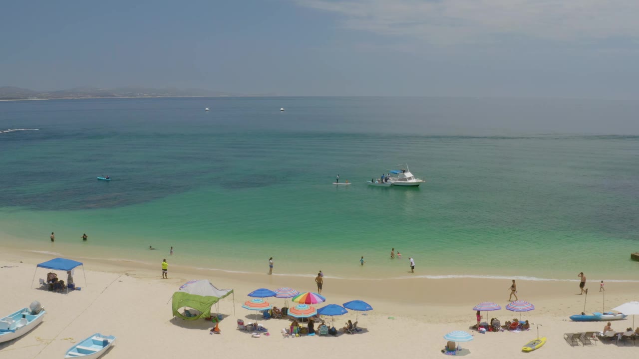 Palmilla Beach aerial frame showing a white boat offshore, darker patches in the green-blue water, umbrellas on the lower beach, and small distant people in the shallows.
