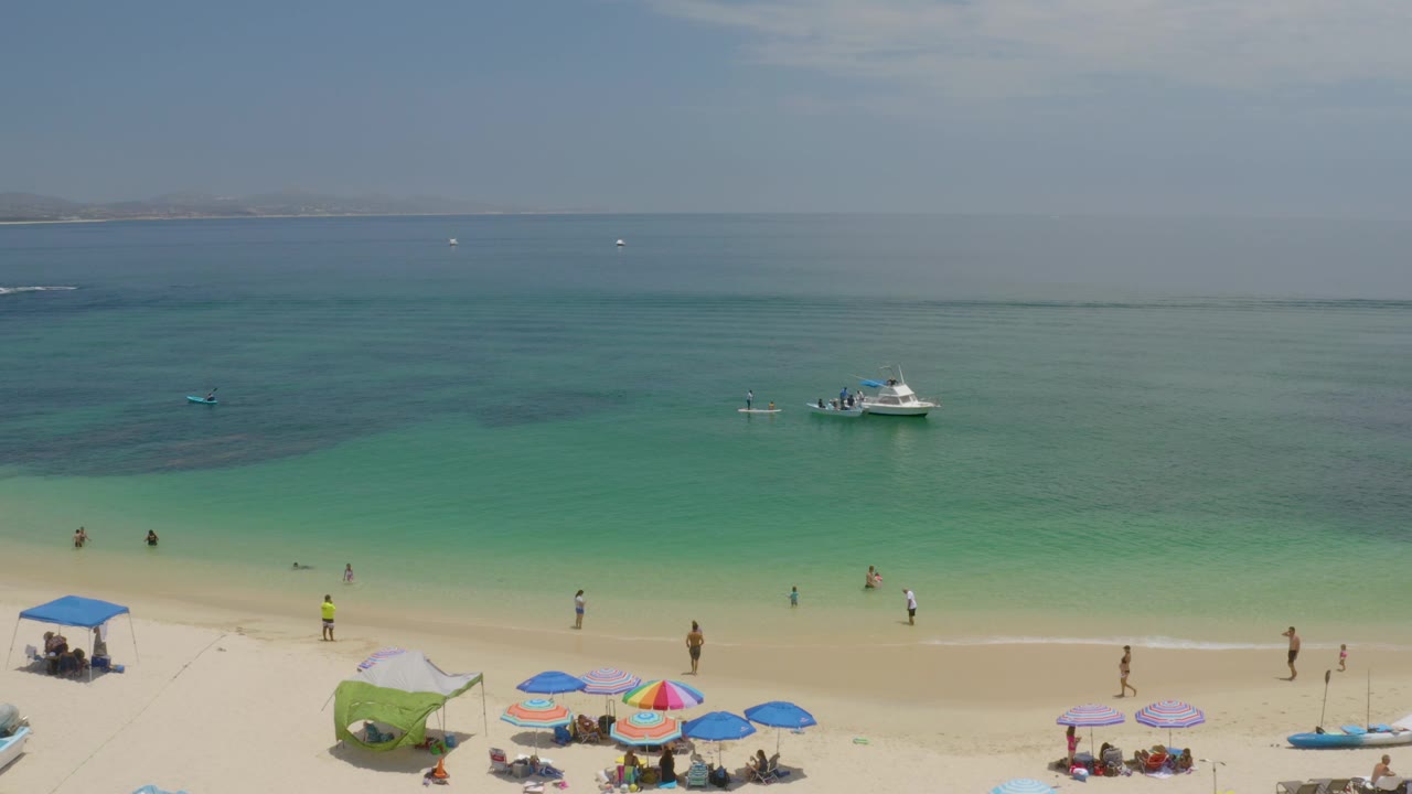 A wide aerial frame over Palmilla Beach with clustered umbrellas at the lower edge, people spread along the shoreline, calm turquoise water, and a small boat offshore.