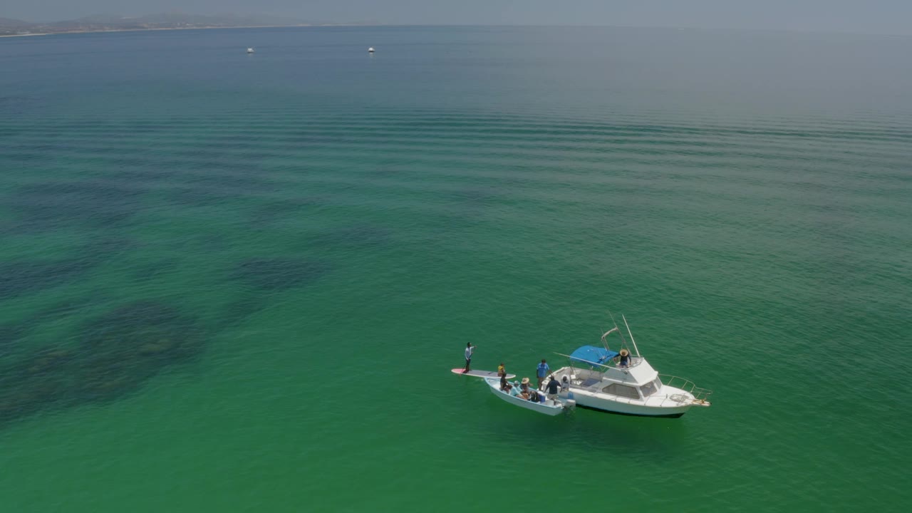 Aerial view of the Palmilla Beach water with the white boat near the right side, people visible aboard, and dark submerged shapes beneath the green surface.