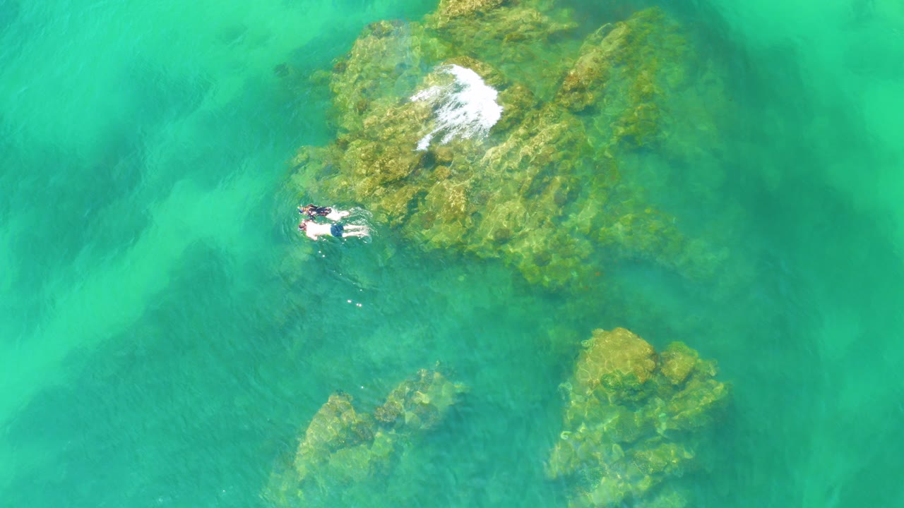 At Palmilla Beach, Baja California Sur, two swimmers appear near the left side of algae-covered reef rocks under clear green water with a small patch of white surf near the upper rock.