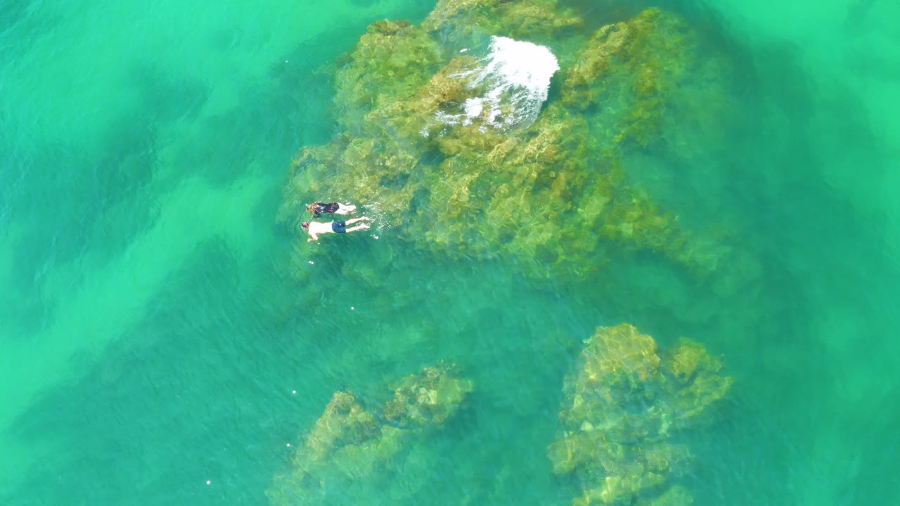 At Palmilla Beach, Baja California Sur, two swimmers are positioned beside the reef as white water breaks across the upper edge of the rocks and darker submerged areas appear below.