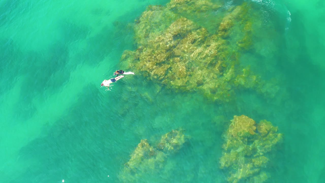 Two snorkelers appear near the left edge of a Palmilla Beach aerial frame, beside a large shallow reef mass in bright turquoise water.