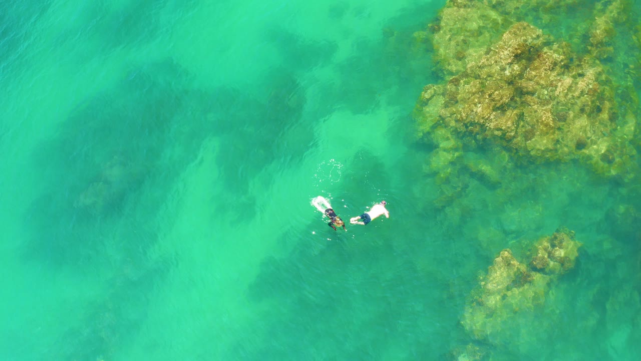 Overhead view at Palmilla Beach, Baja California Sur, showing two swimmers close together in clear turquoise water with rocky reef shapes visible to the right.