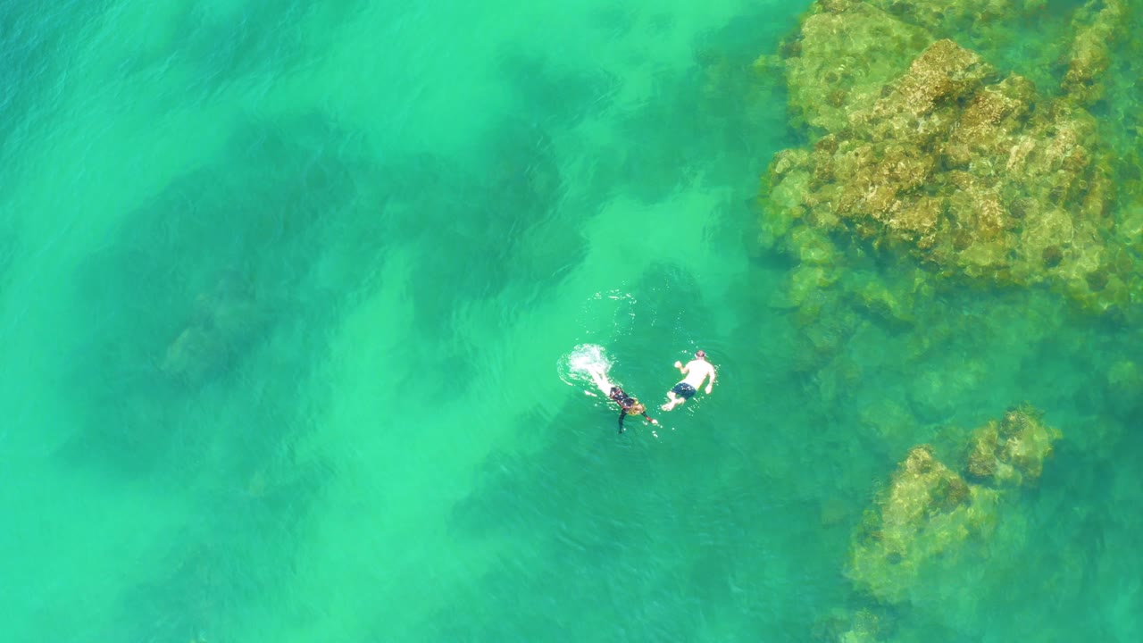 At Palmilla Beach, the overhead view places the swimmers near the right side, with bright surface sparkle, turquoise water, and rocky reef masses above and below them.