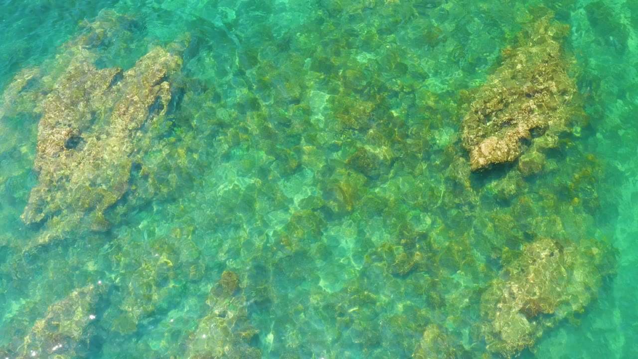 Top-down Palmilla Beach water frame with turquoise ripples, mottled green seabed texture, and two main submerged rock forms, with no visible people.