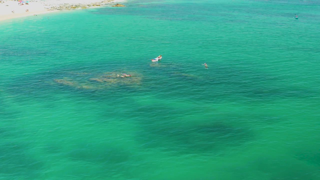Aerial frame over Palmilla Beach, Baja California Sur, showing turquoise water, a pale sand strip at the top, darker underwater rocks, a small white boat, and no visible people.