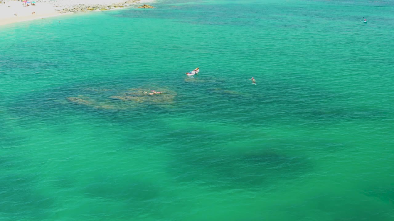 Bird's eye frame from Palmilla Beach with the shoreline along the upper edge, green-blue water across the frame, dark reef patches below the surface, a small white boat, and no visible people.