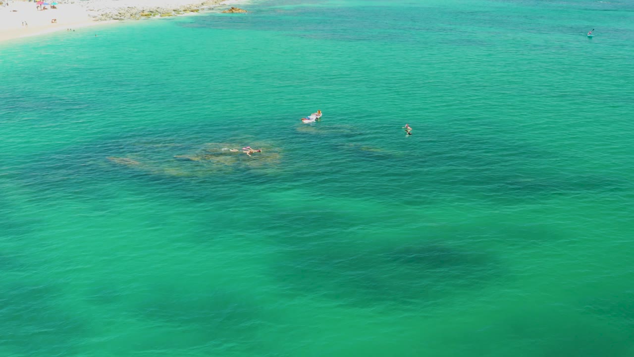 High aerial view at Palmilla Beach showing clear green-blue ocean, a narrow sandy beach edge, soft wave lines, darker underwater patches, a small white boat, and no visible people.