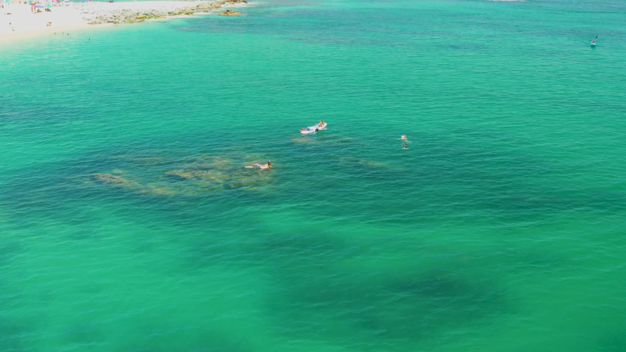 High aerial view over Palmilla Beach, Baja California Sur, with turquoise water, small boats, faint swimmers, sandy shoreline at the top, and distant people too small to identify.