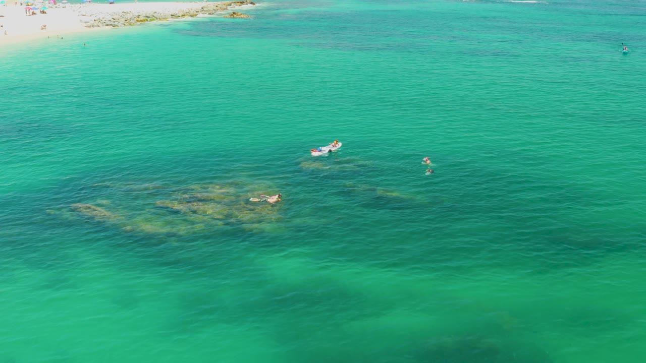 Palmilla Beach aerial with green-blue water filling most of the frame, a small boat and several swimmers set apart from the beach, and darker underwater patches near the middle.