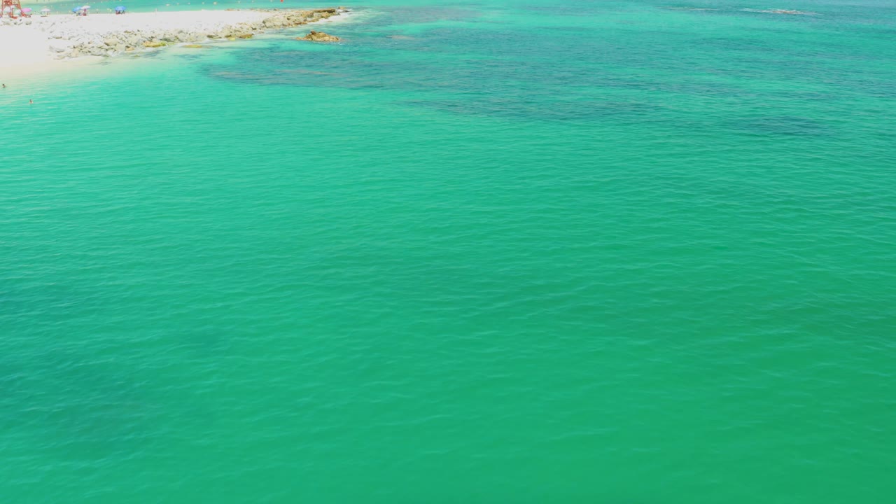 High bird-view frame at Palmilla Beach showing open green-blue water in the foreground, a pale beach strip above, and dark underwater patches with no visible people.