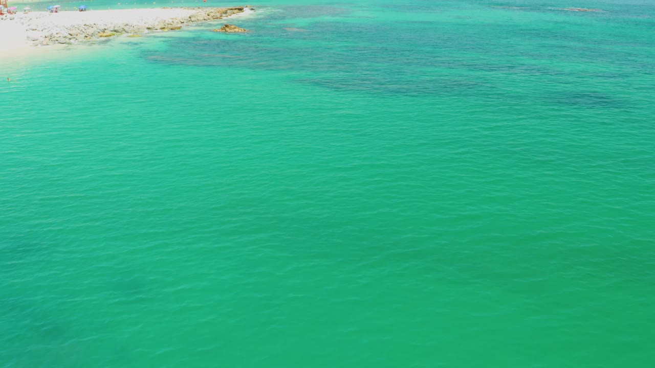 Aerial frame over Palmilla Beach shallows with clear teal water, pale sand at the upper left, and darker reef or rock shapes under the surface, no visible people.