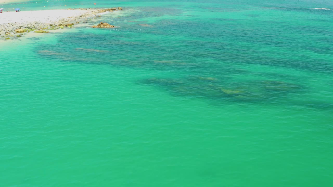 Palmilla Beach aerial frame with turquoise water dominating the image, darker underwater rock or reef areas across the middle, a pale beach strip near the top, and no visible people.