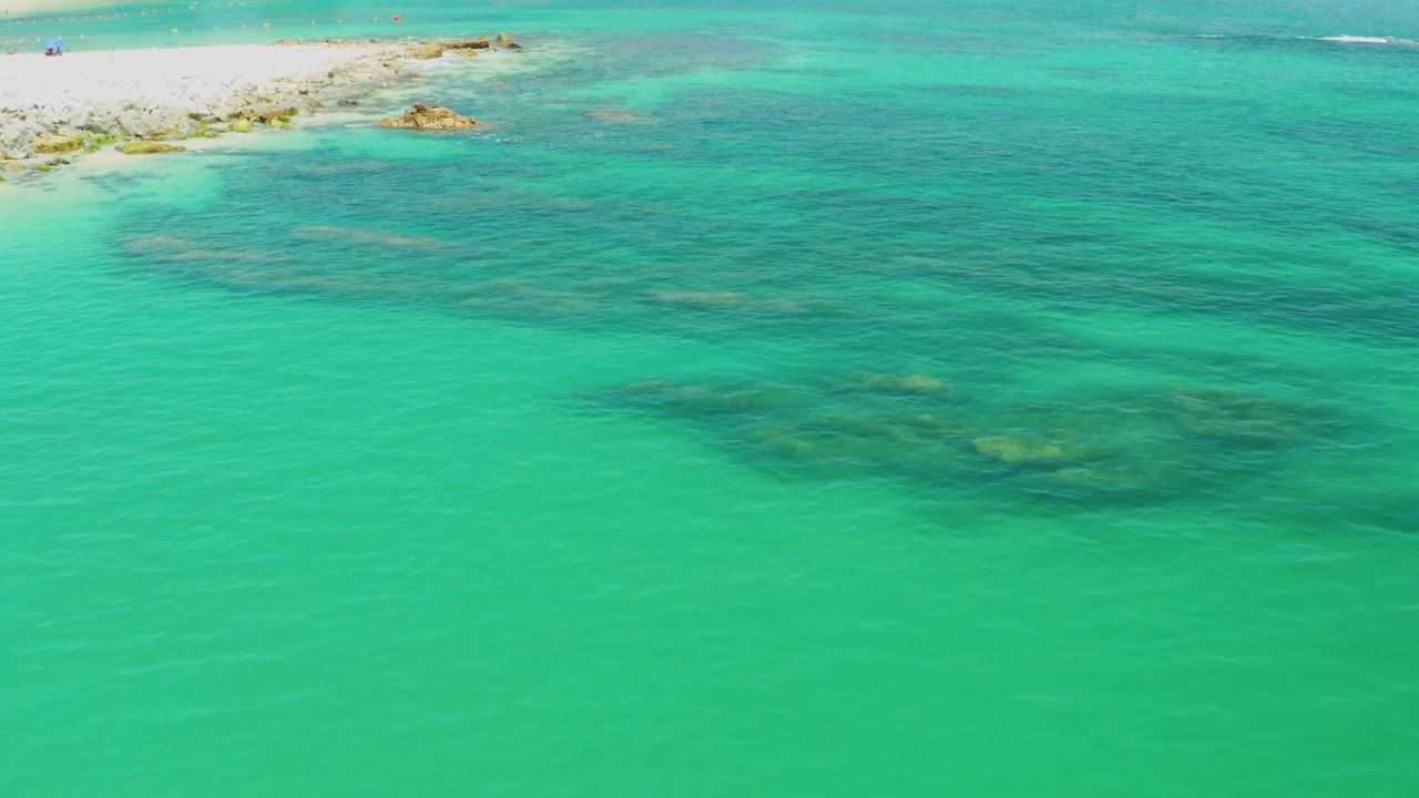 Aerial view at Palmilla Beach with shallow turquoise water, reef shadows spreading across the lower half, pale rocks and sand at the upper left, and no visible people.