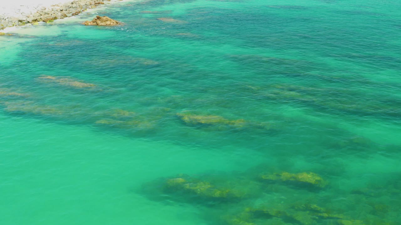 Aerial view over clear turquoise water at Palmilla Beach, Baja California Sur, with dark reef patches below the surface, a small rocky outcrop near the top edge, and no visible people.