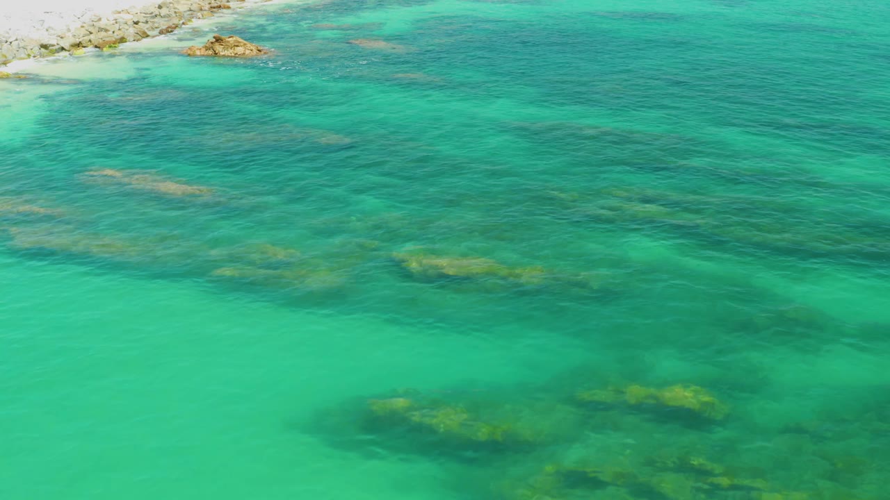 Palmilla Beach aerial frame with clear blue-green water, darker submerged rock or reef shapes across the center, a pale rocky edge at the top, and no visible people.