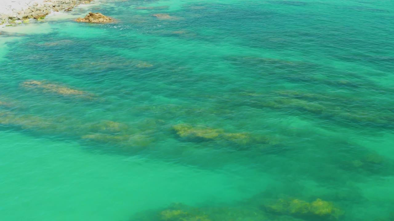 Aerial view at Palmilla Beach showing turquoise water spread across the frame, scattered reef shadows beneath the surface, small exposed rocks near the upper left, and no visible people.