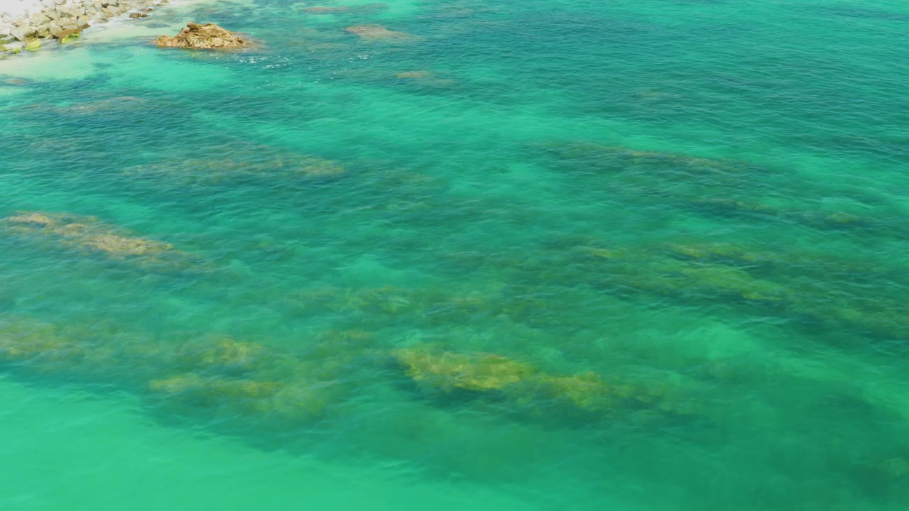 Palmilla Beach water detail with reef bands visible through the turquoise surface, a small rocky outcrop along the upper edge, and no visible people.