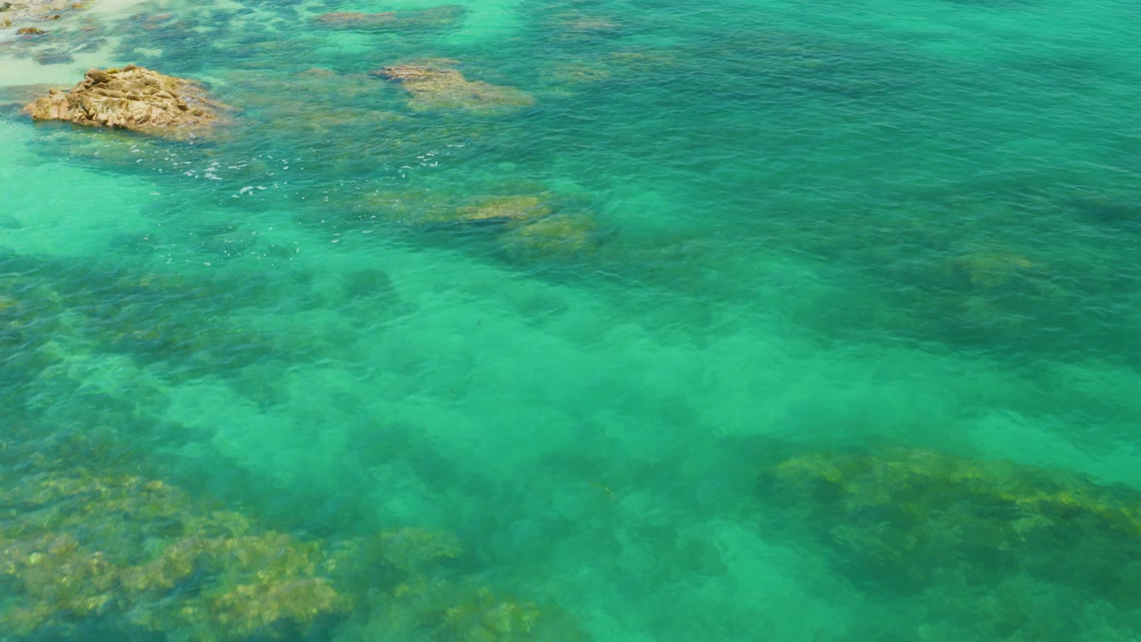 Aerial view over turquoise water at Palmilla Beach, Baja California Sur, with submerged dark rock patches, one tan outcrop near the upper left, and no visible people.