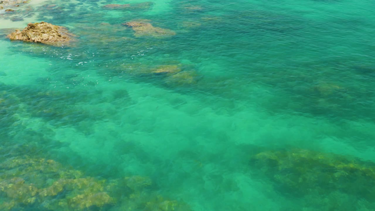 Aerial Palmilla Beach frame showing clear green-blue water, scattered underwater rock forms, a tan rock outcrop near the top edge, and no visible people.