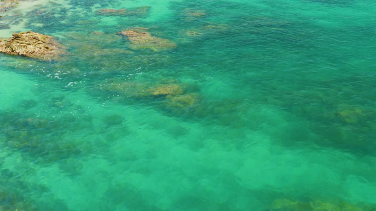 Aerial view at Palmilla Beach with a tan rock outcrop near the upper left, darker submerged patches across the shallow turquoise water, and no visible people.