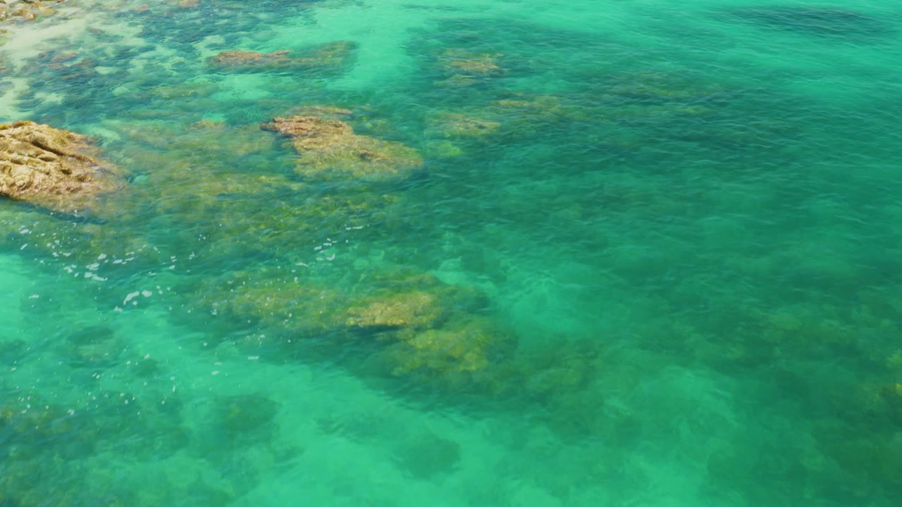 Palmilla Beach aerial frame with rocky ledges entering from the left, transparent blue-green water over darker seabed patches, and no visible people.