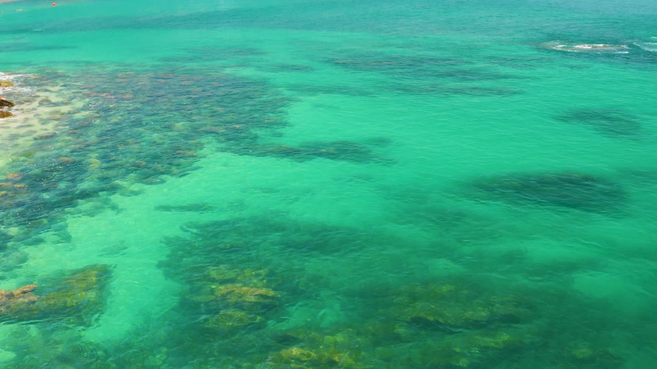 Overhead view of transparent turquoise water at Palmilla Beach, Baja California Sur, with dark submerged formations toward the lower half and no visible people.