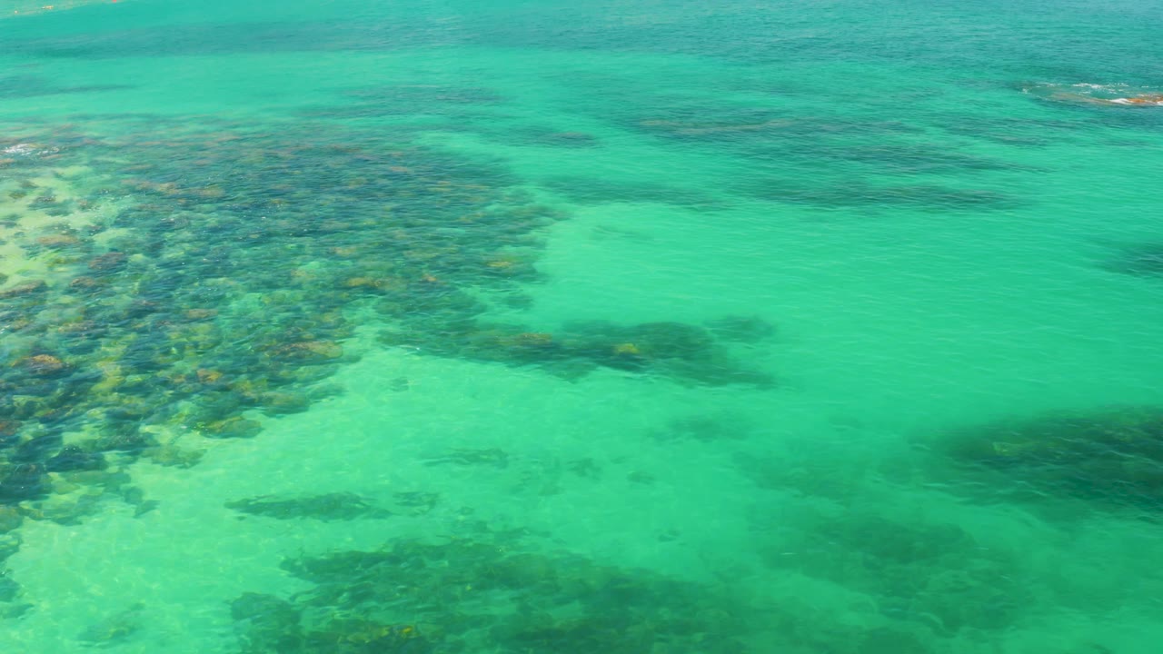 Transparent water at Palmilla Beach reveals mottled rock and sand tones, with the darkest underwater mass moving across the lower right area and no visible people.