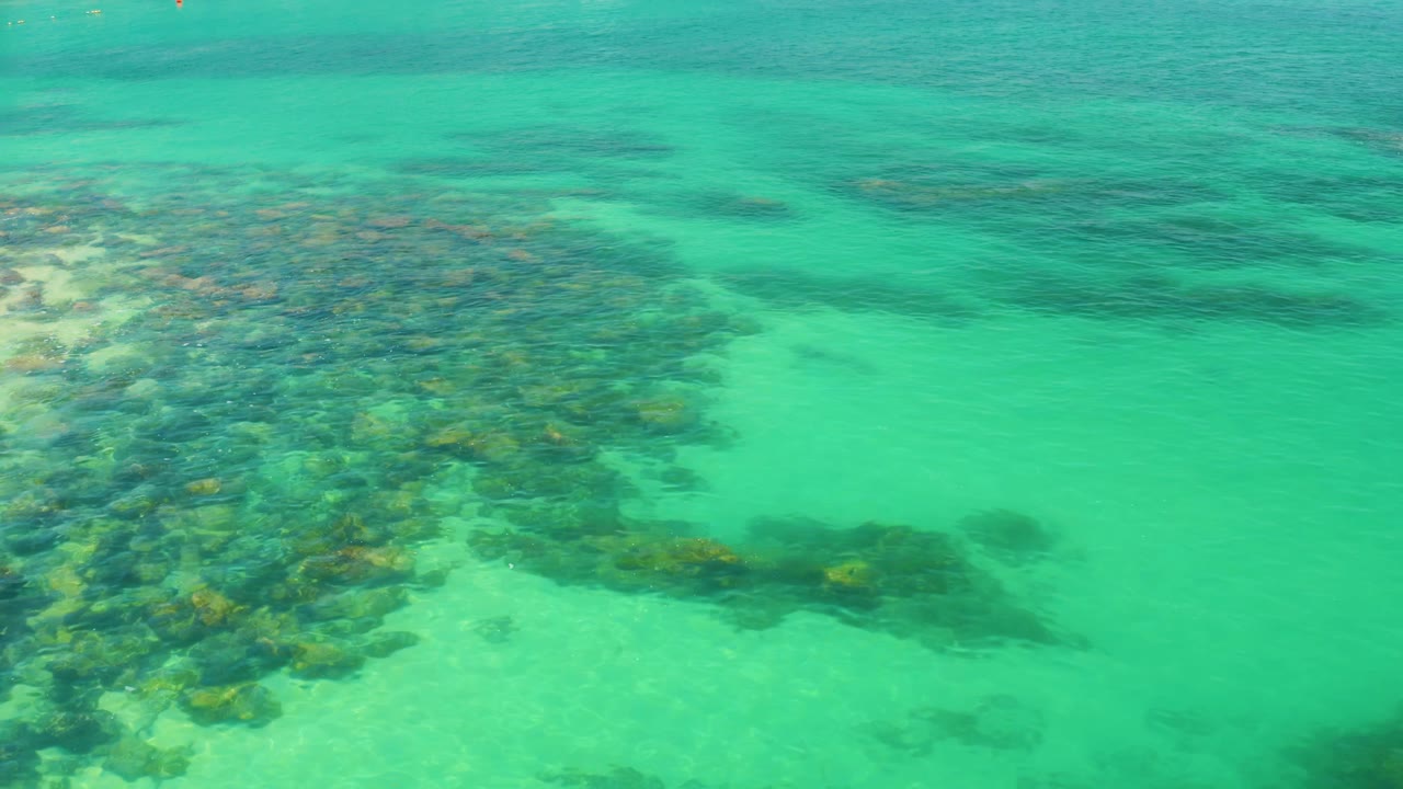 Palmilla Beach shallow water appears green-blue as a broad submerged rock cluster fills the left half of the frame, with lighter sand and no visible people.