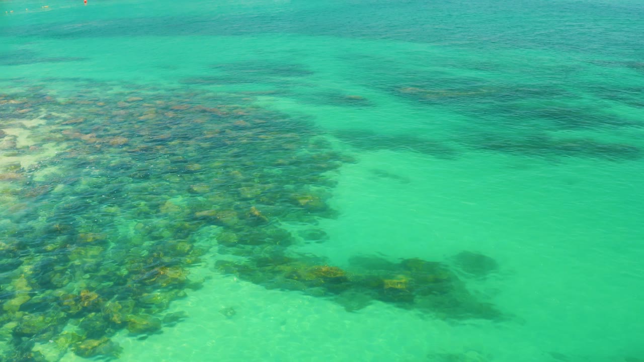 Clear turquoise water at Palmilla Beach reveals overlapping submerged rocks through the lower left and center, with darker underwater patches toward the upper frame and no visible people.