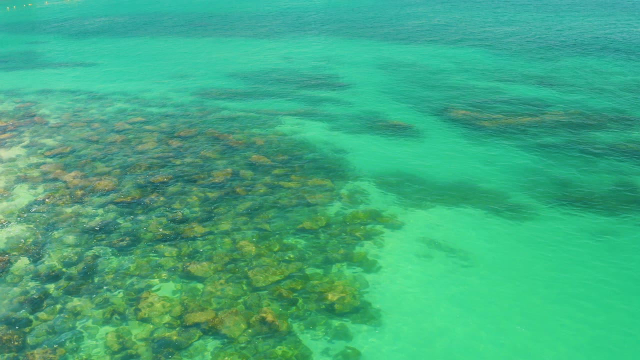 Overhead view of clear green-blue water at Palmilla Beach, Baja California Sur, with reef and sandy bottom visible beneath soft ripples and no visible people.