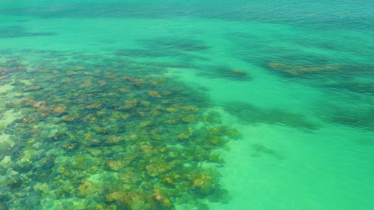 Aerial frame from Palmilla Beach looks down on a dense reef cluster bordered by a turquoise sandy channel, with darker submerged patches in the background and no visible people.