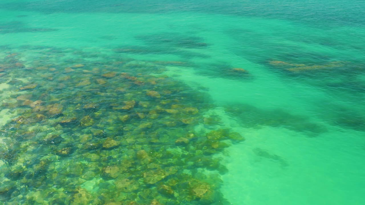 Palmilla Beach shallow water fills the frame with reef along the lower left, pale sand through the center-right, and gentle ripples across the surface with no visible people.