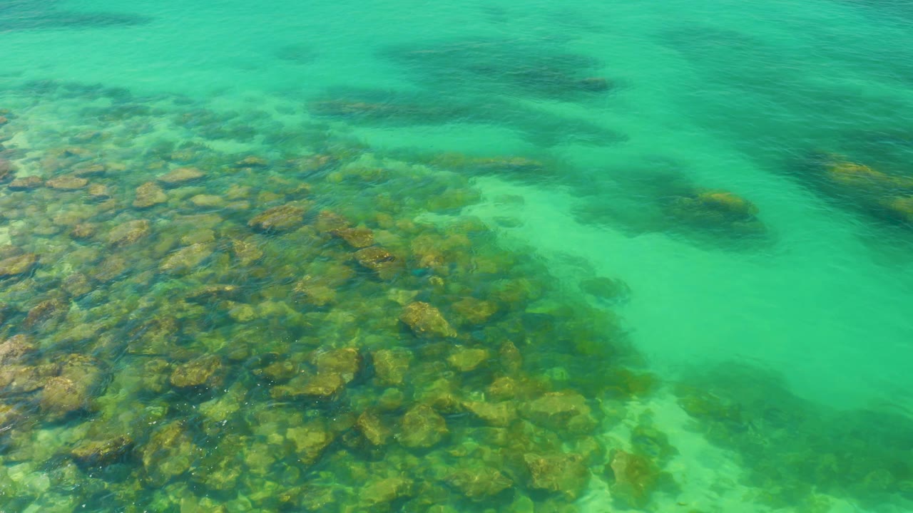 Turquoise shallow water at Palmilla Beach, Baja California Sur, Mexico covers a mixed rocky sea floor, with darker patches farther into the frame and no visible people.