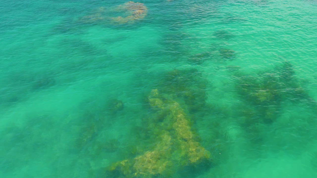 Bird's-eye view of clear green water at Palmilla Beach, Baja California Sur, with submerged rock patches near the center and no visible people.