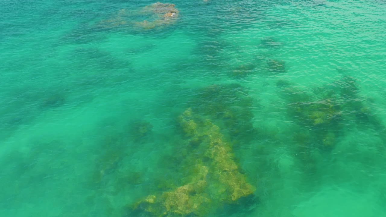Aerial view over Palmilla Beach water, with clustered dark and yellow-green submerged rocks across the lower half and no visible people.