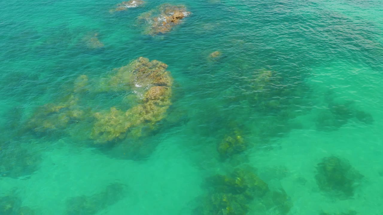 Turquoise shallows cover a curved cluster of rocks at Palmilla Beach, with darker patches beneath the surface and no visible people.