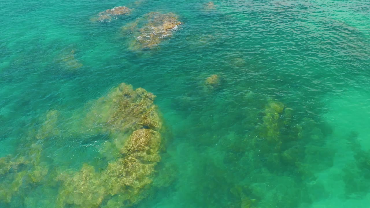 Aerial frame over Palmilla Beach water with a large tan-green submerged rock formation near the lower left and no visible people.