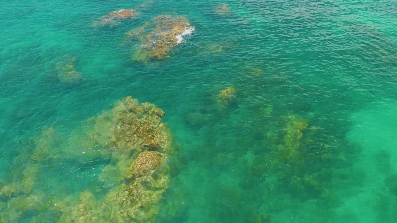 Overhead Palmilla Beach view with a prominent submerged rock mass lower in frame, darker water patches, and no visible people.