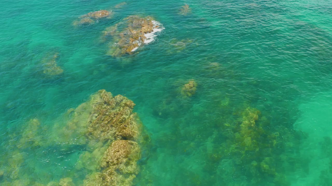 Aerial view at Palmilla Beach with exposed tan rocks near the top, submerged formations below, and no visible people.