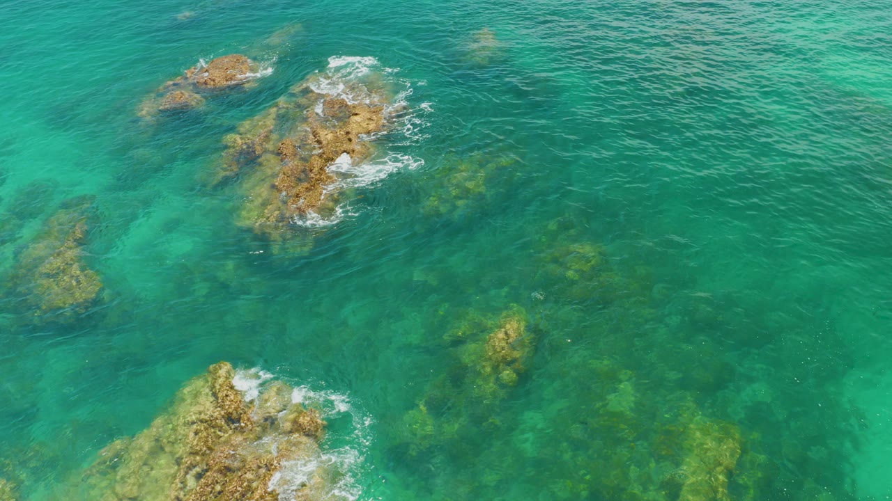 Overhead view of turquoise water at Palmilla Beach, Baja California Sur, with several rocky patches under and above the surface and no visible people.