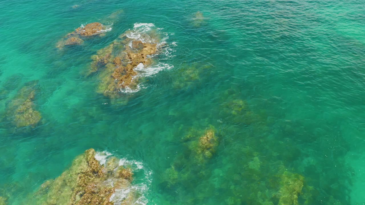 Aerial frame over Palmilla Beach water with exposed rocks near the upper center, submerged rock shapes below, and no visible people.