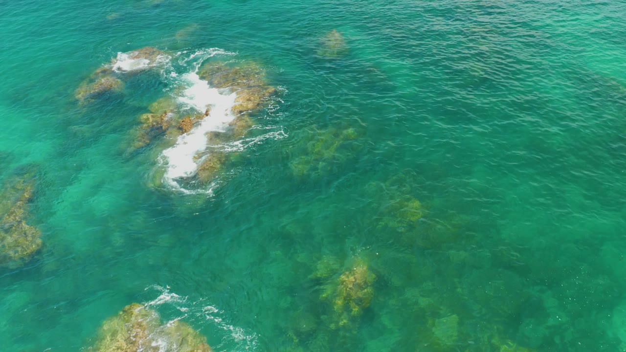 Aerial view of surf breaking harder over a rocky patch at Palmilla Beach, with transparent green water around it and no visible people.