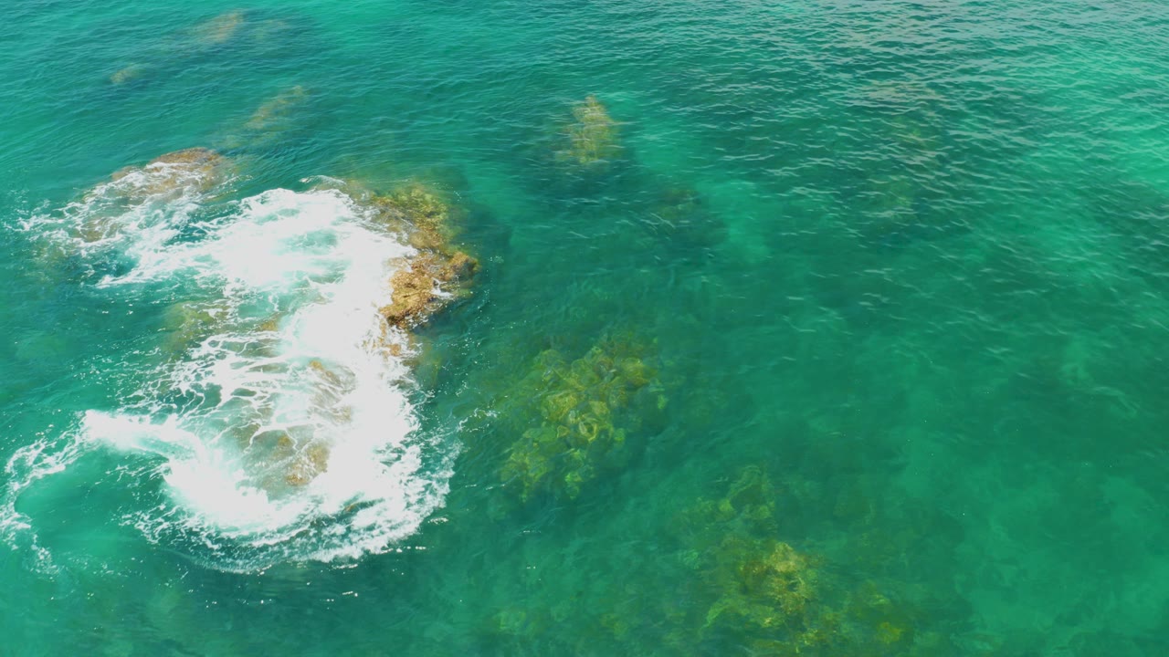 Overhead view at Palmilla Beach, Baja California Sur, with turquoise water and white surf wrapping a tan rock cluster near the left side, no visible people.