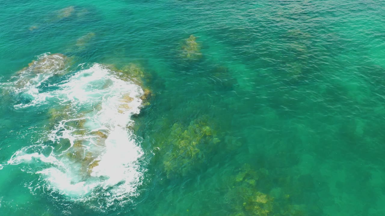 Aerial view at Palmilla Beach with a bright foam line curling around the upper side of the rock cluster and transparent turquoise water filling the frame, no visible people.