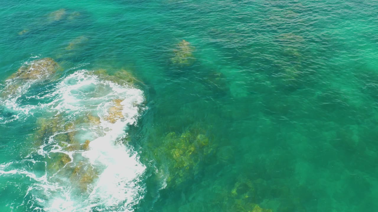 Overhead Palmilla Beach frame where the foamy rock cluster is partly cropped along the left edge, with broad green-blue water and submerged patches extending right, no visible people.