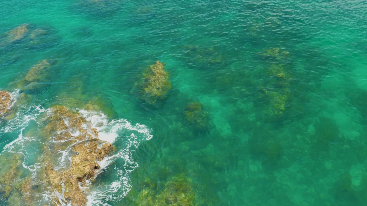 Overhead view at Palmilla Beach showing blue-green water, submerged rock shapes, and white foam breaking around a tan rock at the lower left, with no visible people.
