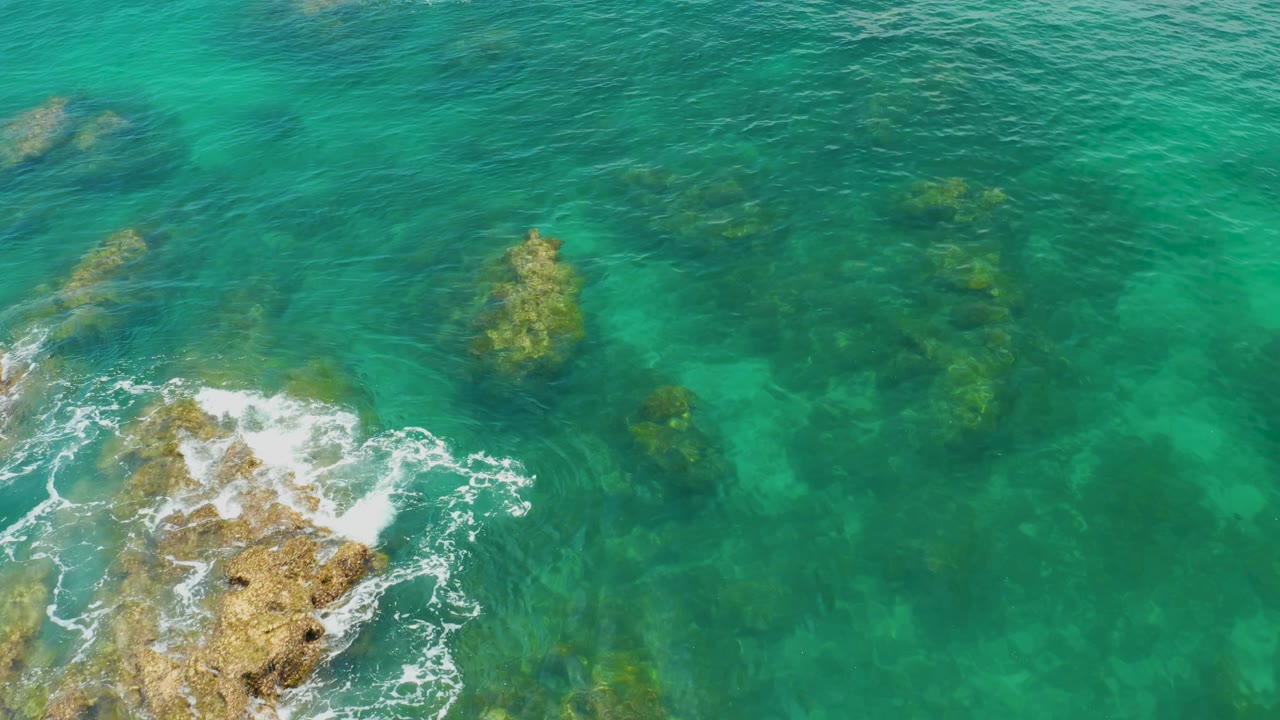 Palmilla Beach water seen from above, with a foamed-over rock crossing the lower edge and darker submerged reef-like shapes in the surrounding blue-green sea, with no visible people.