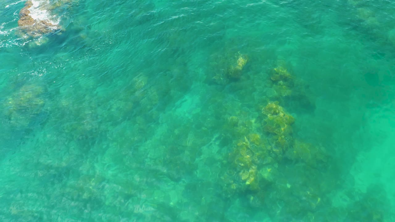 Palmilla Beach water appears green-blue from above, with reef formations visible below and a small foamy rock near the top edge, no visible people.